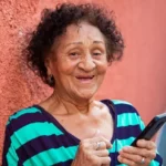 Senhora sorrindo em frente a parede com celular na mão