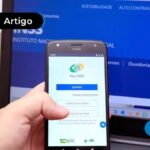 Uma imagem que ilustra um celular na mão em frente a tela do computador com o site do INSS aberto.