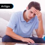 imagem de jovem sentado em sofá com expressão preocupada enquanto usa calculadora