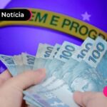 imagem de mão segurando notas de cem reais e ao fundo imagem da bandeira do brasil