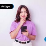 Mulher de cabelos pretos segurando um celular com uma mão ao queixo com expressão de pensativa