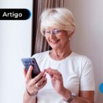 Imagem de idosa com blusa branca segurando celular e sorrindo para a tela