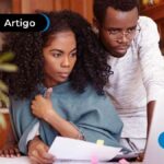 Imagem ilustra jovem casal afrodescendente heterossexual olhando para tela do laptop e com papéis nas mãos.