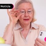 Mulher idosa branca de com cabelos brancos e arco bege na cabeça. veste blusa bege e olha tela de um celular enquanto segura seu óculos com uma mão e o celular com a outra.