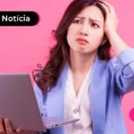 Mulher jovem com cabelos escuros e roupa social segurando notebook com uma mão e a outra mão na cabeça com expressão de confusa