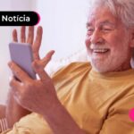 Homem idoso branco de barba e cabelos brancos, com camisa amarela sentado em um sofá, olha a tela de um celular com sorriso no rosto e uma das mãos levantadas em expressão de comemoração.