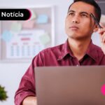 Pessoa com expressão de dúvida vestindo camiseta social vermelho-escuro, sentada em uma mesa de escritório com um notebook aberto. Uma das mãos segura um lápis apontando para sua cabeça enquanto a outra está abaixada encostando na mesa.