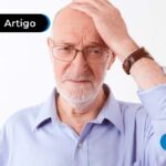 Pessoa de idade usando camiseta social azul-claro e óculos escuro. Com semblante de preocupação ou dúvida, uma das mãos está na cabeça.