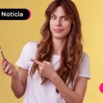 Jovem com cabelo longo solto e blusa branca segurando um celular enquanto aponta para ele com o dedo indicador
