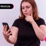 Jovem de cabelo ruivo e camiseta preta segurando um celular com expressão de dúvida.