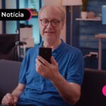 Pessoa idosa com uma camiseta na cor azul marinho, lendo algo no celular com expressão de alegria enquanto está sentado em um sofá.