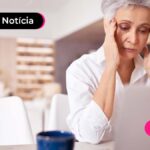 Pessoa idosa vestido uma camiseta com tonalidade clara, olhando para um notebook com as mãos na cabeça sinalizando preocupação.