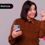 Pessoa idosa de cabelo curto e escuro com expressão de quem teve uma ideia. Vestindo jaqueta marrom enquanto segura um celular e aponta o dedo indicador para cima com a outra mão.