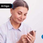 Pessoa idosa com expressão de felicidade, usando camiseta social branca enquanto lê algo em seu celular.