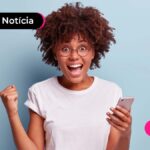 Pessoa de cabelo cacheado e solto, com expressão de felicidade. Uma das mãos está segurando um celular enquanto a outra está fechada e apontando para cima como se estivesse comemorando uma vitória.
