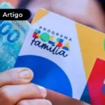 Mão de uma pessoa segurando cartão do Bolsa Família e uma nota de 100 reais na frente do seu rosto.