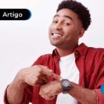 Jovem sorridente apontando para o relógio que está no seu pulso.