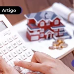 Imagem mostra mão segurando calculadora e miniatura de casa no fundo.