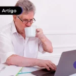 Pessoa de idade segurando xícara branca e mexendo no computador.