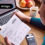 Pessoa segurando boleto, ao lado um notebook aberto e calculadora.