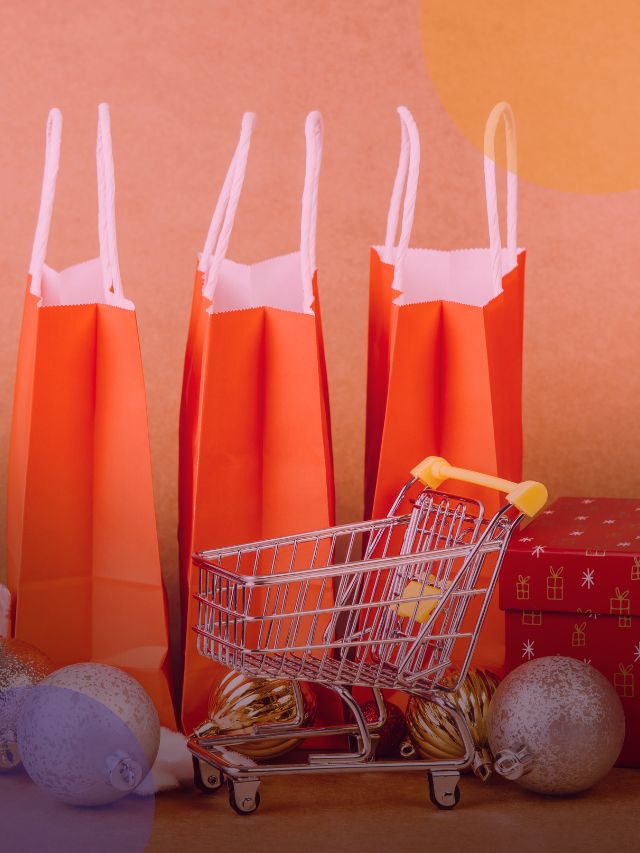 Sacolas de presente vermelhas e mini carrinho de compras com decoração de natal ao lado.
