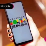 Mão de uma pessoa com unhas pintadas de preto segurando um celular que aparece a logo do Programa Bolsa Família na tela.
