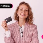 Pessoa de roupa social com óculos pendurado na camisa e segurando um cartão enquanto sorri para câmera.