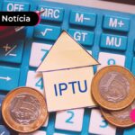 Calculadora azul sobre um caderno aberto, com caneta, e moedas espalhadas acima, e um papel em formato de casinha escrito "IPTU".