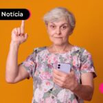 Pessoa idosa de cabelos brancos curtos com o celular em uma mão abaixo do rosto, e outra mão apontando para cima.