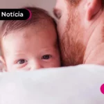 Pessoa de costas com camisa branca segurando um bebê no colo com a cabeça em seus ombros.