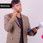 Pessoa usando um blazer marrom segurando uma prancheta numa mão e caneta na outra encostando-a na cabeça.