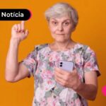 Pessoa de mais idade com celular em uma mão e apontando pra cima com a outra.