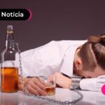 Pessoa sentada com a cabeça baixa sobre a mesa, acorrentada com uma garrafa e segurando um copo.