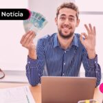 Pessoa de camisa social e óculos segurando notas de dinheiro em uma mão e fazendo sinal de "OK" com a outra, sentado em uma cadeira de frente para notebook e papéis.