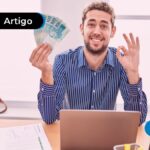 Pessoa sorrindo segurando notas de cem reais, em frente a um computador