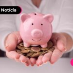 Mão segurando cofrinho rosa e moedas.