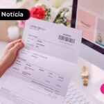 Mão feminina segurando um boleto impresso, com uma monitor e mesa de escritório ao fundo.