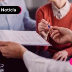 Pessoa com terno assinando documento e outras duas pessoas ao lado.