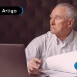 Pessoa de idade com expressão de dúvida, segurando papel branco e com computador na mesa.