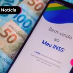 Celular com aplicativo do INSS e notas de cinquenta e cem reais ao lado.