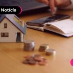 Pessoa no celular calculando o IPTU representado por uma casa de brinquedo e pilhas de moedas sobre a mesa