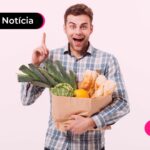 Jovem sorrindo enquanto segura sacola de mercado e ler notícia sobre queda dos preços dos alimentos.