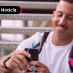 Pessoa de blusa branca e mochila segurando o celular enquanto está sorrindo.