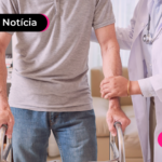 Pessoa de camisa cinza usando um andador enquanto pessoa de jaleco auxilia.