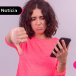 Pessoa de blusa rosa segurando celular com a mão esquerda enquanto aponta o dedo para baixo com a mão direita.