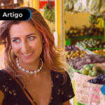 Imagem mostra uma mulher sorrindo em uma feira, destacando frutas e verduras ao fundo, ilustrando tema de economia e compras conscientes.