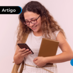 Jovem estudante sorridente, usando óculos e vestido branco, segura livros e olha para o celular enquanto caminha.