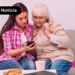 Idosa sentada em um sofá com uma mulher, olhando para o celular.