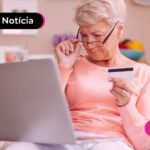 Idosa olhando para um notebook e segurando um cartão de crédito.