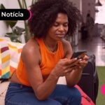 Uma mulher de camiseta laranja sentada olhando para o celular.